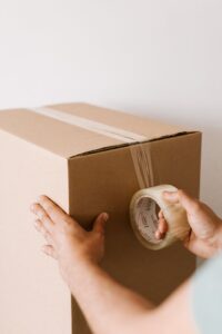 Crop anonymous male using tape to seal packed cardboard box against white wall while moving into new place